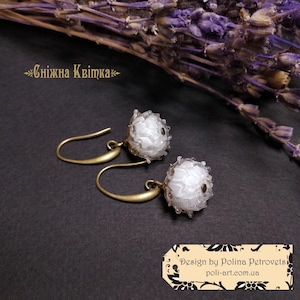 Könnte beinhalten: Ein Paar weiße Blumenohrringe mit goldfarbenen Haken. Die Ohrringe haben ein zartes, strukturiertes Aussehen, mit kleinen, klaren Perlen um die weiße Blume. Der Hintergrund ist dunkel, mit getrocknetem Lavendel und dem Text "Сніжна Квітка".
