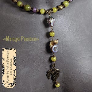May include: A handcrafted necklace featuring a pendant with a seashell, leaf, and bead accents. The necklace includes green and purple beads, with the text "Design by Polina Petrovets" on a tag.