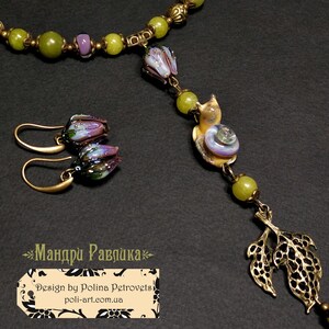 May include: A handcrafted jewelry set featuring a necklace and earrings. The necklace has green beads, a snail pendant, and a leaf charm. The earrings have purple and green flower-shaped beads. The set is made with brass-colored metal.