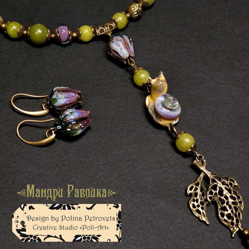 May include: A handcrafted jewelry set featuring a necklace and earrings. The necklace has green beads, a snail pendant, and a leaf charm. The earrings have purple and blue flower-shaped beads. The set is made with gold-colored metal.
