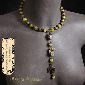 May include: A beaded necklace with green and purple beads. The pendant features a cat, a pearl, and a decorative leaf. The necklace is displayed on a mannequin.