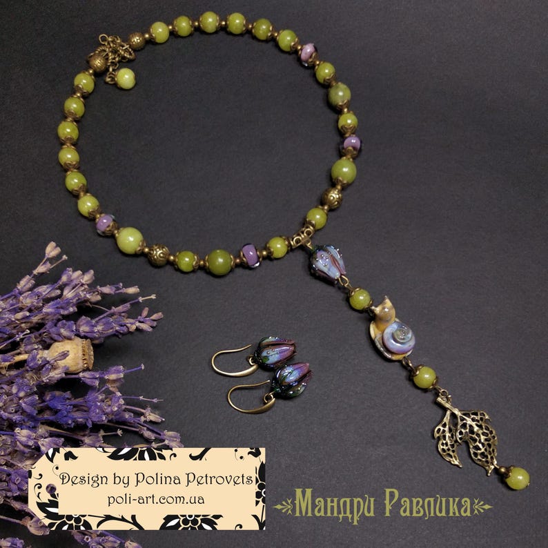 May include: A handmade jewelry set featuring a necklace and earrings. The necklace is composed of green and purple beads, with a pendant of a snail and leaf. The earrings have a similar design. The set is displayed on a dark surface.