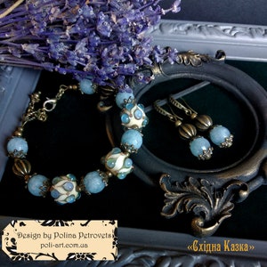 Puede incluir: Un conjunto de pulsera y pendientes de cuentas azules y blancas con detalles en bronce. La joyería se muestra sobre un fondo de terciopelo negro con un marco decorativo. El texto "Design by Polina Petrovels poli-art.com.ua" y "<Східна Казка>" es visible en la imagen.