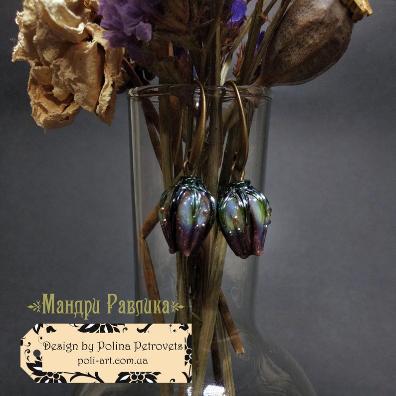 May include: A pair of iridescent, handcrafted earrings with a floral design. The earrings feature a dark green and purple color scheme with gold-colored ear wires. The earrings are displayed in a clear glass vase with dried flowers.