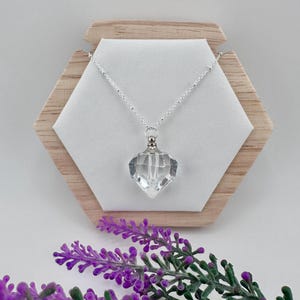 May include: A clear crystal pendant necklace with a faceted, diamond-shaped pendant. The pendant hangs from a delicate silver chain. The necklace is displayed on a white surface against a wooden hexagonal backdrop, with purple flowers in the foreground.