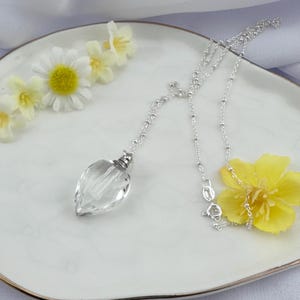 Può includere: Una collana a catena d'argento con un ciondolo di cristallo trasparente a forma di lacrima. La collana è su un piatto bianco con fiori gialli.