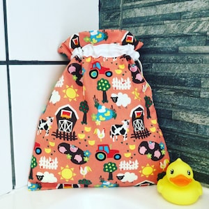 Puede incluir: Una bolsa de cordón hecha de tela naranja con un ribete blanco. La bolsa presenta un estampado de temática agrícola con imágenes de graneros, tractores, vacas, cerdos, pollos y ovejas. La bolsa está sentada sobre una superficie de azulejos junto a un pato de goma amarillo.
