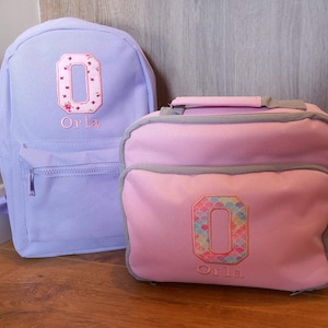 May include: A purple backpack and a pink lunch bag with the letter "O" embroidered on each. Both bags have the name "Orla" embroidered below the letter.