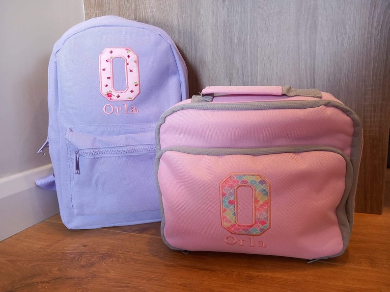 May include: A purple backpack and a pink lunch bag with the letter "O" embroidered on each. Both bags have the name "Orla" embroidered below the letter.