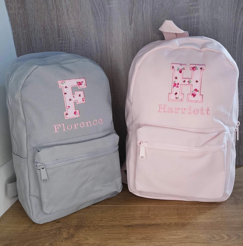 May include: Two personalized backpacks, one gray with the letter F and the name Florence embroidered on it, and one pink with the letter H and the name Harriett embroidered on it.