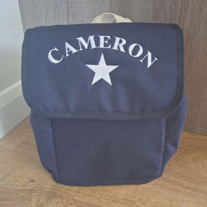 Peut inclure: Sac bleu marine avec fermeture à rabat, de style sac à dos. Le mot "CAMERON" est brodé en blanc au-dessus d'une étoile blanche. Il possède une poignée beige. Le sac est posé sur une surface en bois.