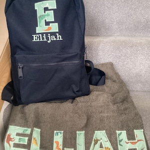 May include: A black backpack with a green embroidered letter E and the name Elijah. A green towel with the name Elijah embroidered in green letters with a dinosaur pattern.