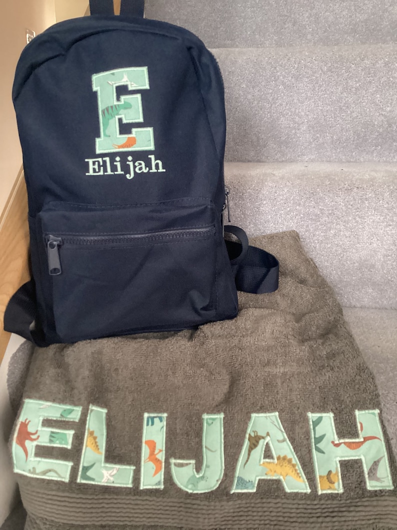 May include: A black backpack with a green embroidered letter E and the name Elijah. A green towel with the name Elijah embroidered in green letters with a dinosaur pattern.