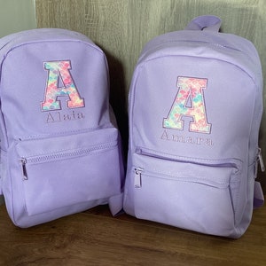 May include: Two purple backpacks with a large letter A on the front. The letter A is decorated with a mermaid scale pattern in pink, blue, and green. The backpacks have the names Alaia and Amara embroidered below the letter A.
