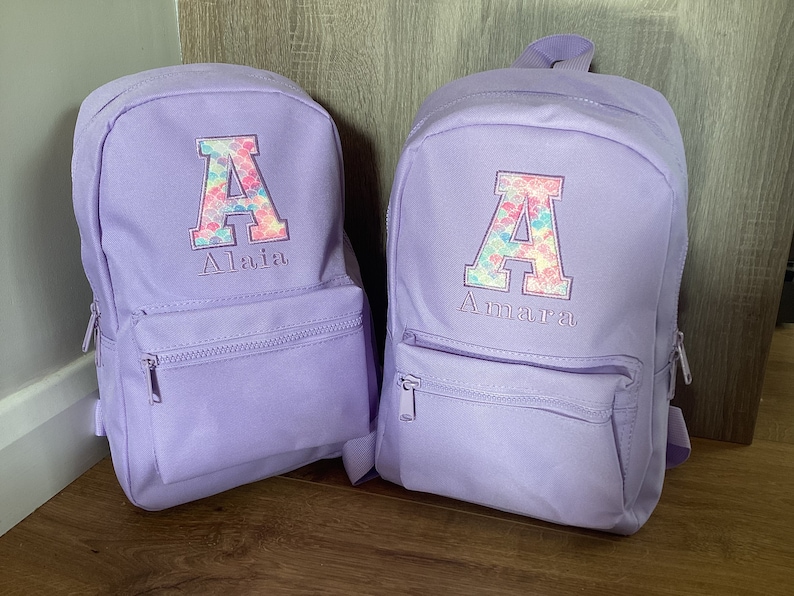 May include: Two purple backpacks with a large letter A on the front. The letter A is decorated with a mermaid scale pattern in pink, blue, and green. The backpacks have the names Alaia and Amara embroidered below the letter A.