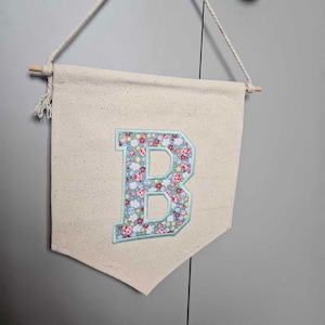 May include: A white canvas banner with a floral patterned letter "B" in the centre. The banner is hanging from a rope and has wooden dowels at the top and bottom.