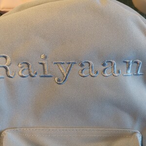 May include: A light blue backpack with the name "Raiyaan" embroidered in white on the front.