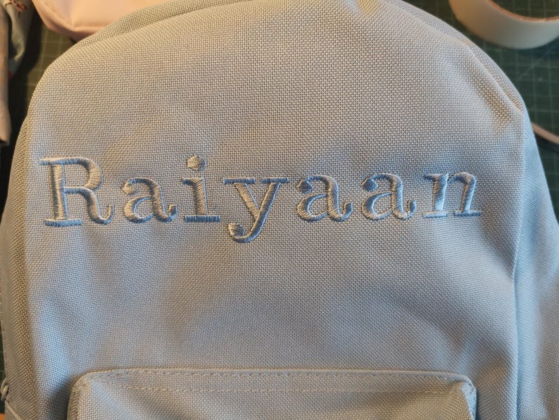 May include: A light blue backpack with the name "Raiyaan" embroidered in white on the front.
