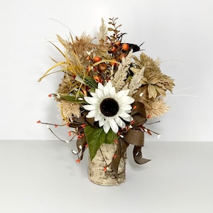 May include: A decorative floral arrangement featuring a large white sunflower with a dark center, surrounded by dried flowers, berries, and greenery. The arrangement is set in a birch bark vase and accented with a brown ribbon.