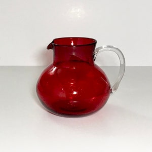 May include: A red glass pitcher with a clear glass handle. The pitcher has a round body and a wide mouth.