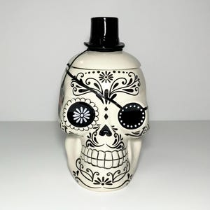 May include: A ceramic skull-shaped jar with a black top hat lid. The off-white jar is decorated with black floral and swirl designs, an eye patch, and a flower-shaped eye. The jar has a wide mouth and a detailed jawline.