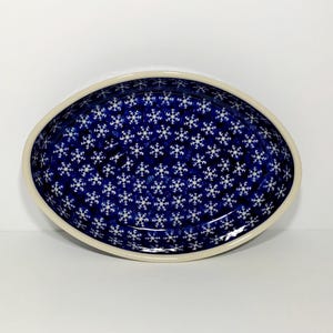 May include: Oval ceramic serving dish with a dark blue interior decorated with white snowflake patterns. The dish has a cream-colored rim and is set against a white background. A decorative piece for serving food.
