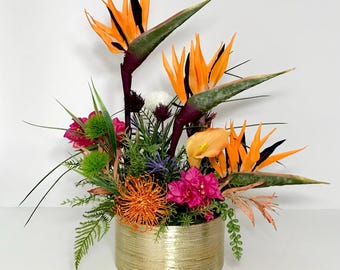 Puede incluir: Un arreglo floral tropical en un jarrón dorado. El arreglo presenta flores de ave del paraíso naranja, buganvillas rosas, protea de cojín naranja y follaje verde.