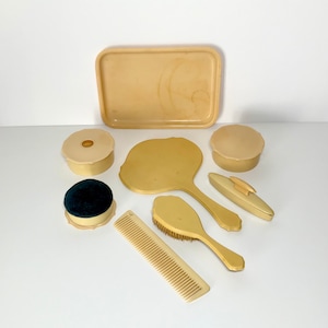 May include: A vintage vanity set in a cream color, including a tray, two round containers, a hand mirror, a brush, a comb, and a puff.