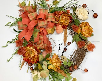 May include: A fall-themed wreath with a rustic grapevine base. It features orange, yellow, and cream-colored flowers, accented with green foliage, berries, and a large orange plaid bow. A decorative piece for the autumn season.