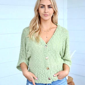 May include: A light green, button-up cardigan with a v-neck and a slightly cropped length. The cardigan is made of a soft, textured fabric and has a relaxed fit.