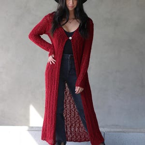 May include: A long, red, open-front cardigan with a single button closure. The cardigan is made of a lightweight, knit fabric and has a relaxed fit. The model is wearing the cardigan over a black tank top and dark wash jeans.