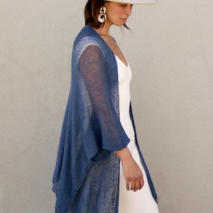 May include: A sheer, open-front blue cardigan with a loose, draped fit and three-quarter length sleeves, worn over a solid white sleeveless top and white trousers, paired with a wide-brimmed cream-colored hat featuring a metallic band.