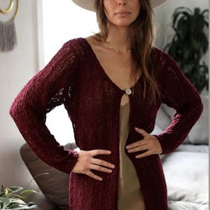 May include: A long, burgundy cardigan with a single button closure. The cardigan is made of a lightweight, open-weave fabric and has a relaxed fit.