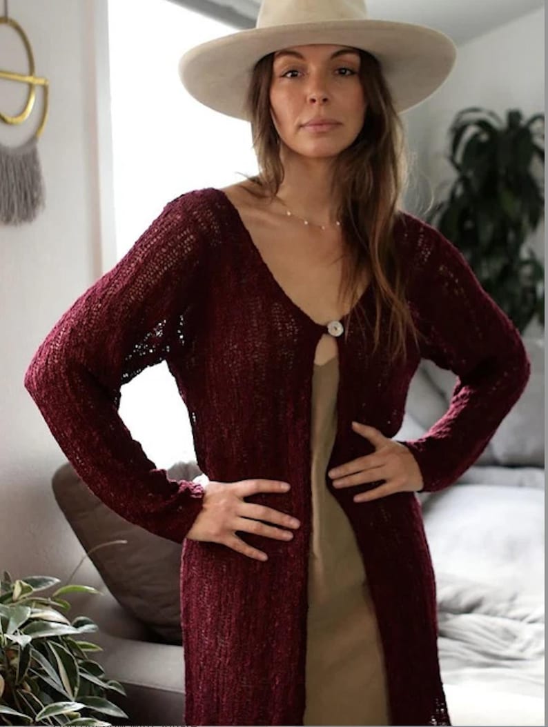 May include: A long, burgundy cardigan with a single button closure. The cardigan is made of a lightweight, open-weave fabric and has a relaxed fit.