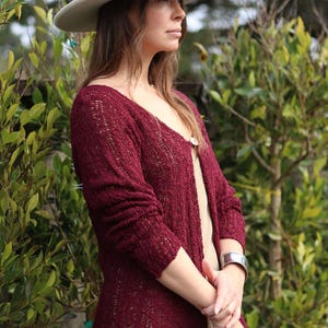 May include: A woman wearing a burgundy knit cardigan with a white hat. The cardigan has a loose fit and is open in the front. The woman is standing in front of a green leafy background.