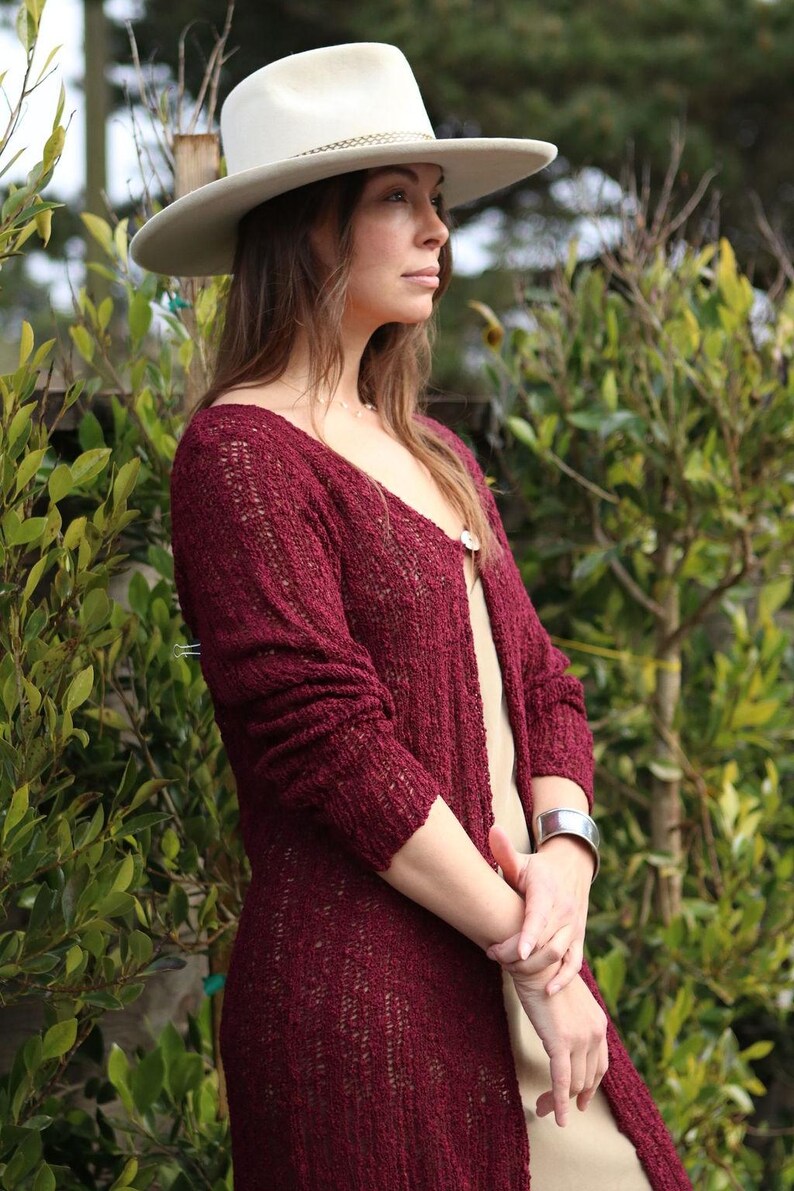 May include: A woman wearing a burgundy knit cardigan with a white hat. The cardigan has a loose fit and is open in the front. The woman is standing in front of a green leafy background.