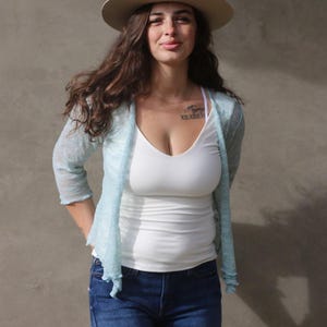 Può includere: Una donna indossa un cardigan blu chiaro sopra una canotta bianca e jeans blu. Indossa un cappello fedora beige. La donna ha i capelli lunghi e castani e sorride.