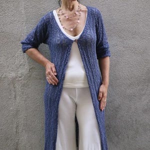 May include: A woman wearing a blue knit cardigan with a white top and white pants. She is wearing a layered necklace with pink beads.