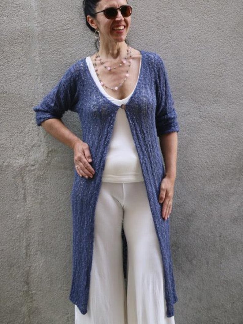 May include: A woman wearing a blue knit cardigan with a white top and white pants. She is wearing a layered necklace with pink beads.