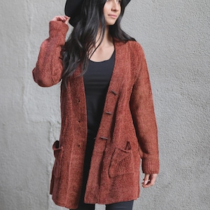 May include: A rust-colored, long-sleeved, textured knit cardigan featuring a button-front closure with toggle-style buttons and two front patch pockets. The garment is worn over a black top and paired with a black wide-brimmed hat.