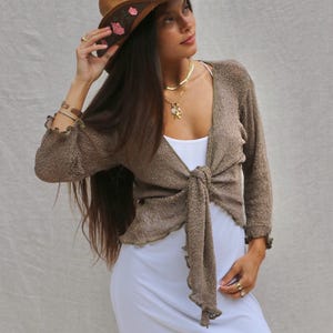 May include: A brown knit cardigan with three-quarter length sleeves and a tie-front closure, worn over a plain white sleeveless dress. The cardigan features a textured fabric with ruffled edges at the cuffs and hem, paired with a brown straw cowboy hat.