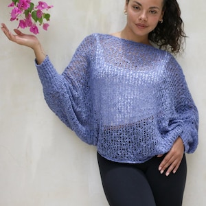 May include: A periwinkle blue loose-knit sweater with a wide boat neckline and long, batwing-style sleeves. The garment features an open, airy crochet-like texture and is worn over a solid white camisole, paired with plain black leggings.