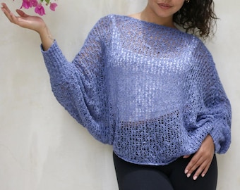 May include: A periwinkle blue loose-knit sweater with a wide boat neckline and long, batwing-style sleeves. The garment features an open, airy crochet-like texture and is worn over a solid white camisole, paired with plain black leggings.