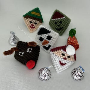 May include: A selection of handmade, diamond-shaped Christmas decorations. The decorations feature festive designs including an elf, a snowman, Santa Claus, and a reindeer. Hershey's Kisses and a strawberry sweet are also present. The text "HUGS" is visible on one of the sweets.