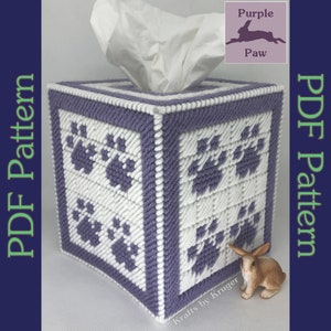 May include: Purple and white tissue box cover with paw prints. The cover is made with plastic canvas and yarn. The cover is sitting on a table with a small rabbit figurine.