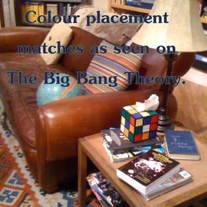 Op de afbeelding: Een bruine leren bank met een gestreept kussen en een globe op de zitting. Een houten bijzettafel met een stapel boeken en een Rubik's Cube-weefselhouder. De tekst "Colour placement matches as seen on The Big Bang Theory." is zichtbaar in de afbeelding.