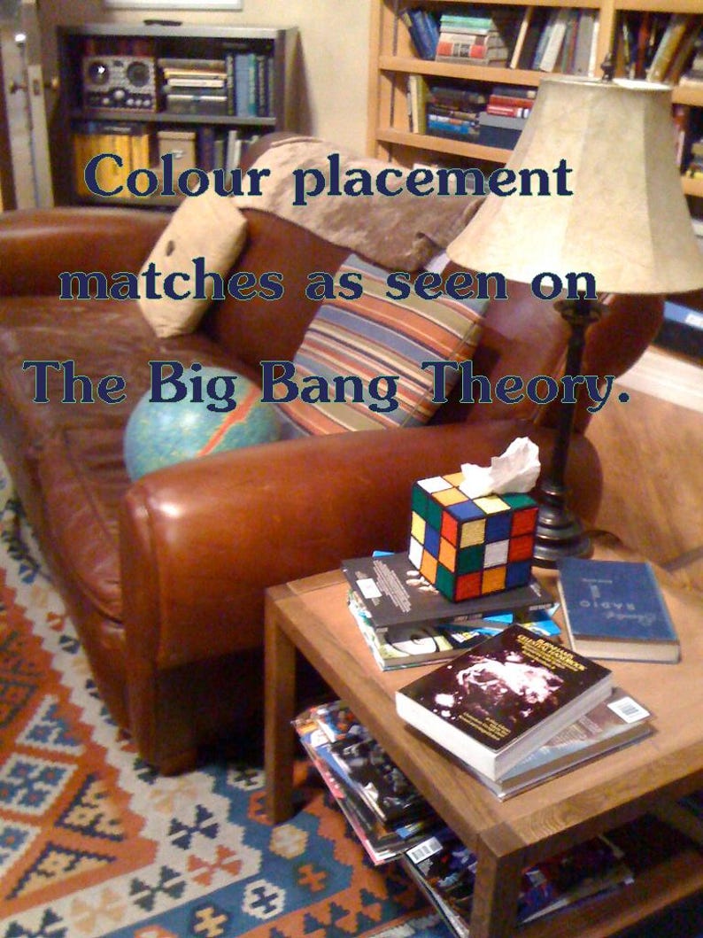 Op de afbeelding: Een bruine leren bank met een gestreept kussen en een globe op de zitting. Een houten bijzettafel met een stapel boeken en een Rubik's Cube-weefselhouder. De tekst "Colour placement matches as seen on The Big Bang Theory." is zichtbaar in de afbeelding.