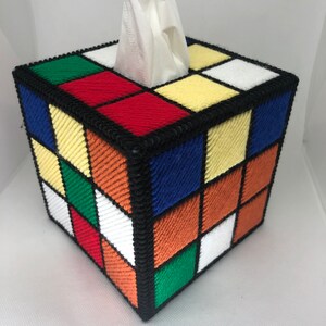 Op de afbeelding: Een tissueboxhoes in de vorm van een Rubik's Cube met een wit papiertje dat uit de bovenkant steekt. De kubus is gemaakt van kleurrijke garenvierkanten in rood, geel, blauw, groen, oranje en wit.