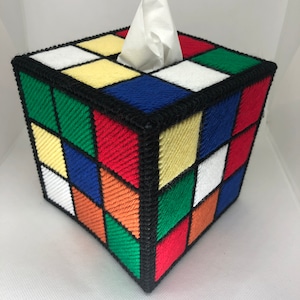 May include: A tissue box cover shaped like a Rubik's Cube with a white tissue peeking out of the top opening. The cube is made of colourful yarn squares in red, green, blue, yellow, orange, and white.
