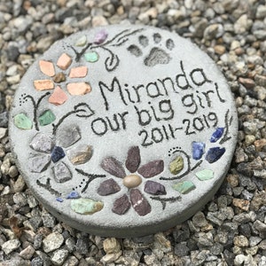 May include: A round, gray stone memorial with the inscription "Miranda our big girl 2011-2019." The stone is decorated with colorful stones and floral designs, including a paw print. The stone is set on a bed of small gray and tan pebbles.